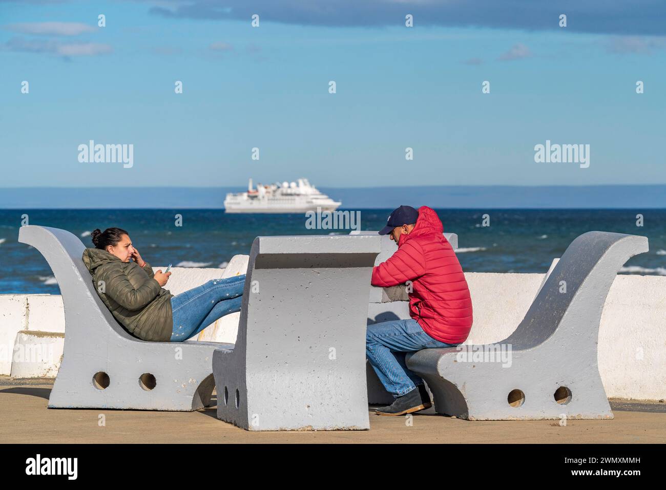 Le persone con abiti invernali caldi siedono sulle panchine sulle rive dello stretto di Magellano, città di Punta Arenas, Patagonia, Cile Foto Stock