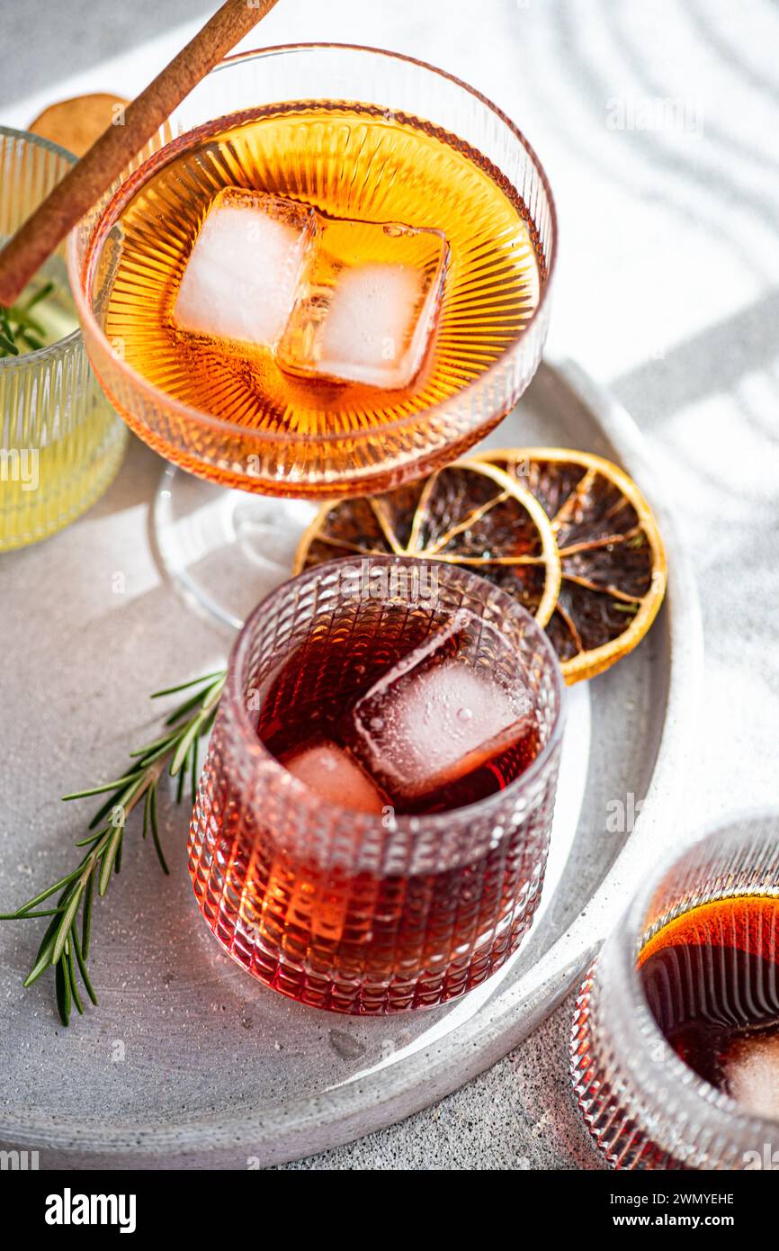 Una serie di squisite bevande alcoliche in bicchieri decorativi, accompagnate da cubetti di ghiaccio, un bastoncino di cannella, rosmarino e fette di agrumi secchi su un ma Foto Stock