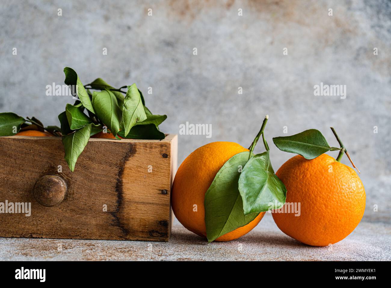 Sono esposte arance mature con foglie verdi fresche attaccate, alcune appoggiate su una superficie ruvida e altre nascoste in una cassa di legno, che evocano un senso Foto Stock