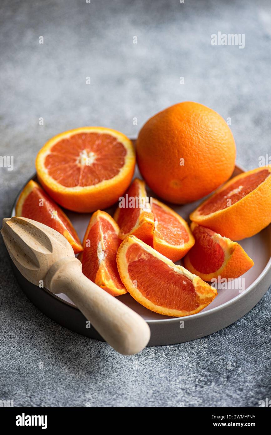 Un piatto pieno di arance mature e tagliate di recente accanto a una centrifuga di agrumi in legno su sfondo grigio testurizzato. Foto Stock
