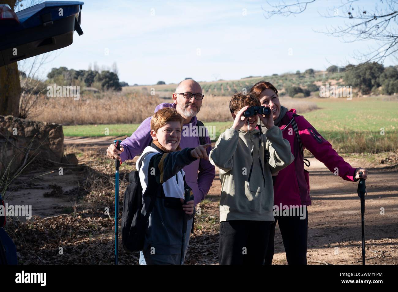 Una famiglia allegra che si gode una giornata di sole facendo escursioni in campagna, con due bambini sorridenti, uno con pali da trekking e l'altro con la binocula Foto Stock