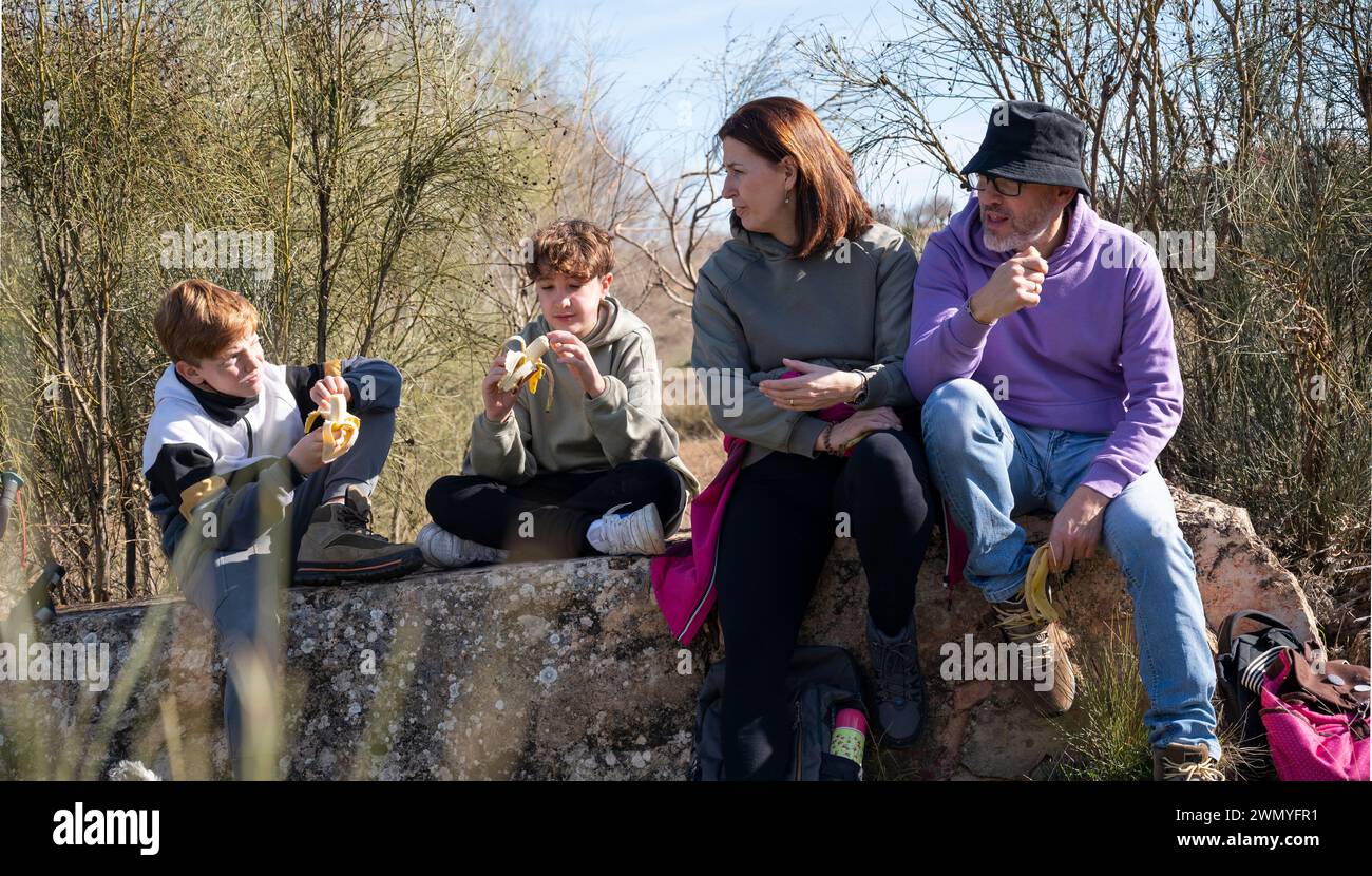 Una famiglia si gode una pausa snack su una panchina di pietra rustica durante un'escursione, con due bambini che sbucciano le banane e gli adulti che guardano Foto Stock