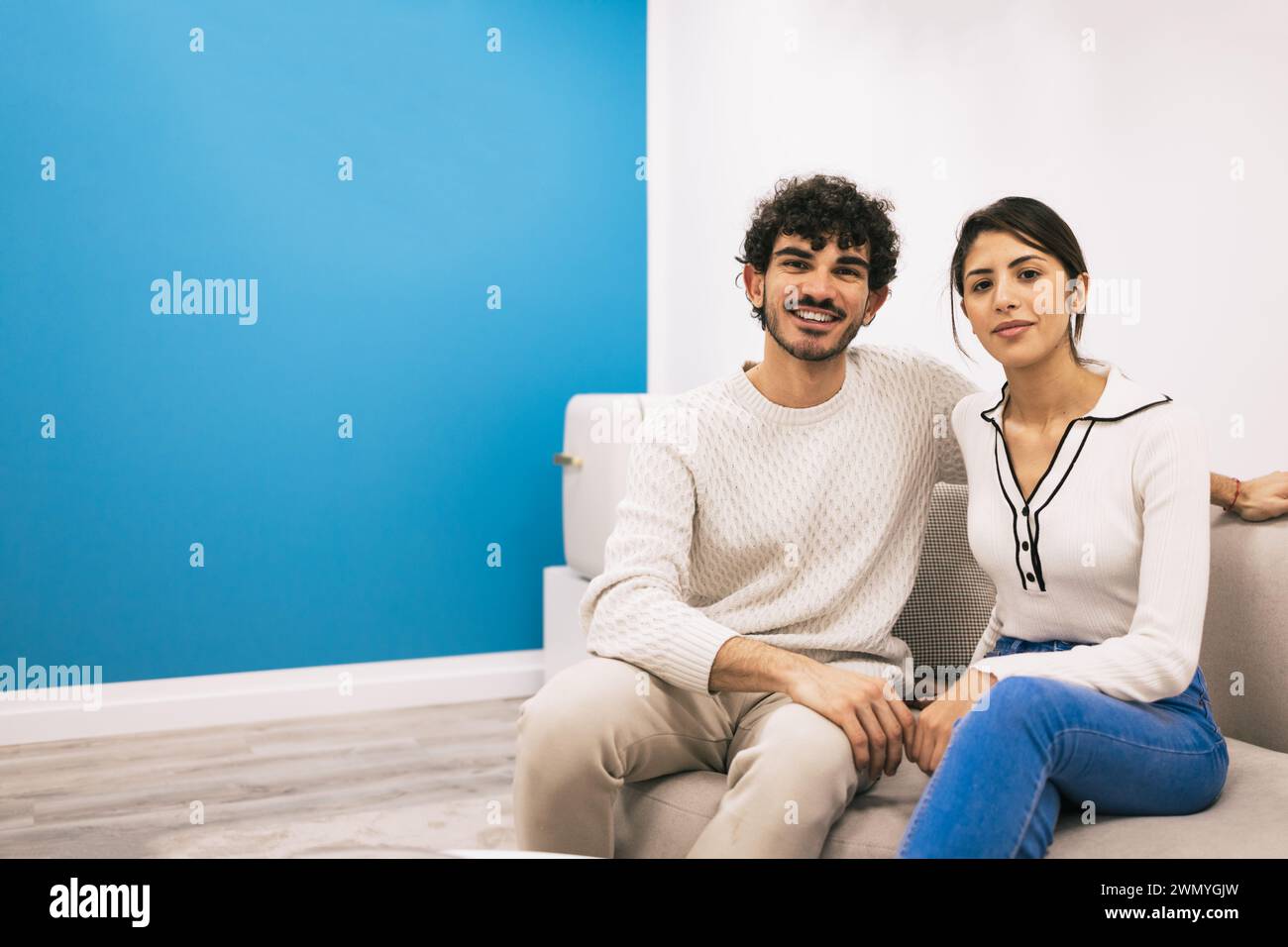Una giovane coppia siede comodamente su un divano bianco, sorridendo e posando casualmente contro un muro blu nella loro accogliente casa. Foto Stock