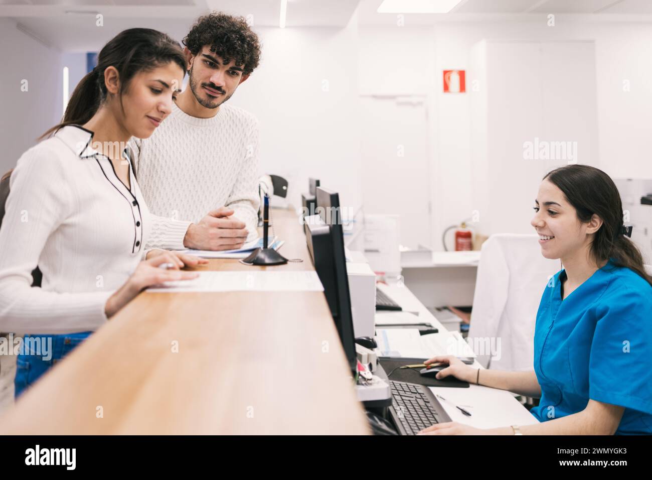 Un cordiale receptionist assiste una coppia alla reception di una clinica medica, fornendo informazioni e indicazioni con un sorriso. Foto Stock