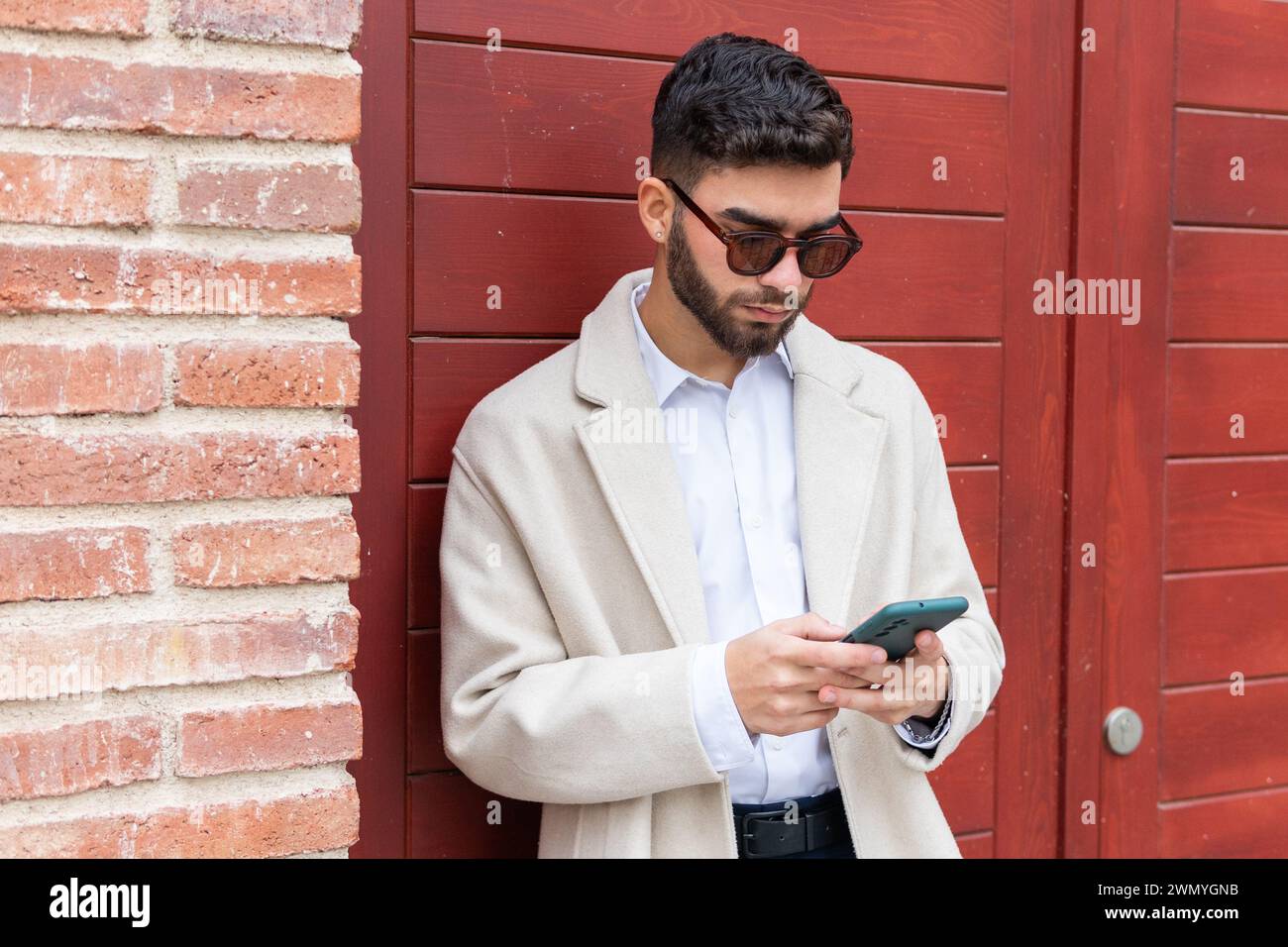 Uomo d'affari alla moda con occhiali da sole, che si concentra sul suo smartphone, accanto a una porta rossa con sfondo in mattoni Foto Stock