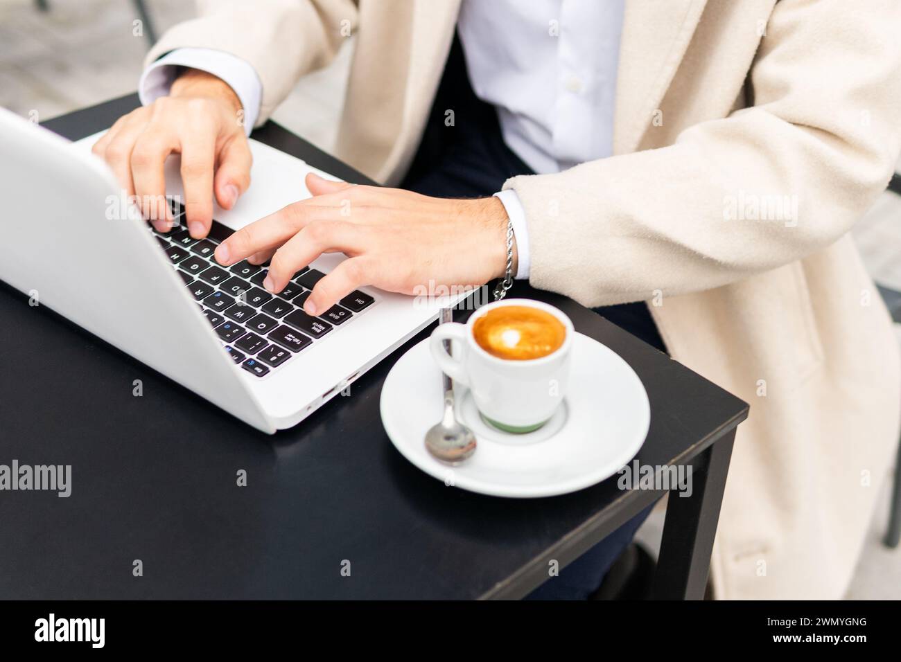 Primo piano di un uomo d'affari anonimo che digita un laptop con un espresso fresco sul tavolo, simboleggiando la produttività e il lavoro in movimento Foto Stock