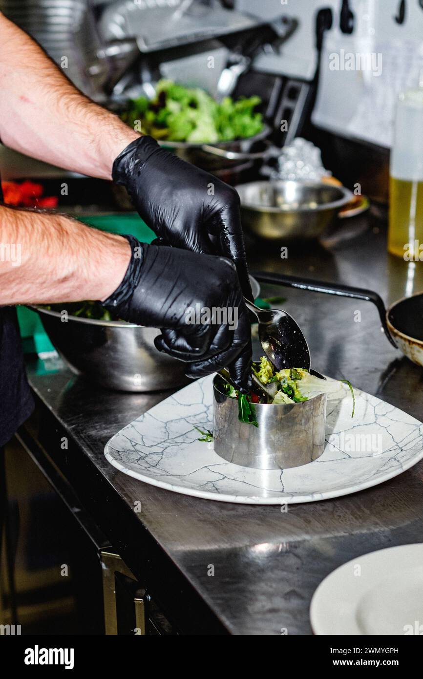 Primo piano delle mani di uno chef con guanti neri che ripiano delicatamente un'insalata gourmet in un anello di metallo su un piatto con motivi di marmo in una cucina professionale Foto Stock
