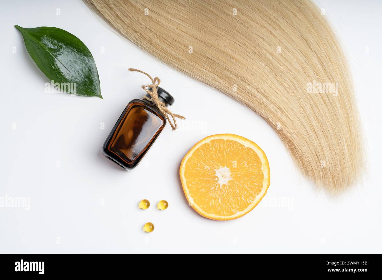 Una ciocca di capelli biondi insieme a prodotti naturali per la cura dei capelli, tra cui una bottiglia di vetro marrone, fetta di arancia, foglia verde e capsule vitaminiche. Foto Stock