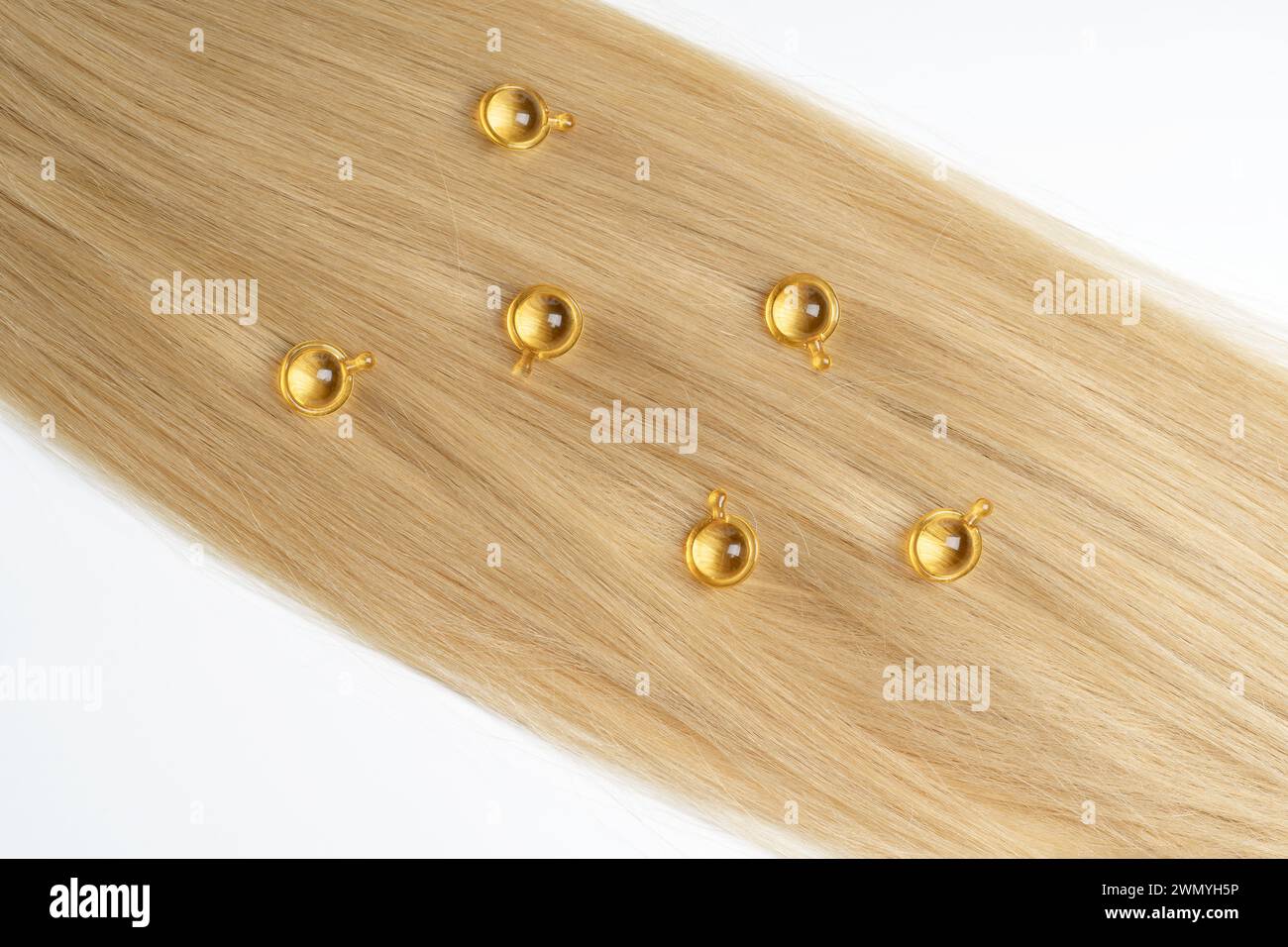 Lussuose capsule dorate per la cura dei capelli posizionate strategicamente su un'estensione liscia dei capelli biondi, che simboleggia la bellezza e il trattamento dei capelli. Foto Stock