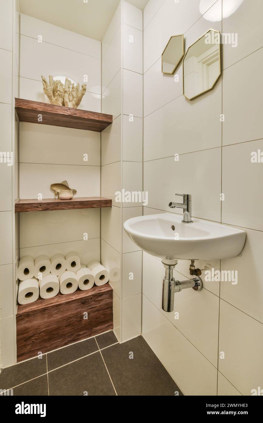 Un piccolo bagno contemporaneo con piastrelle in ceramica bianca, scaffalature in legno, un lavandino in porcellana bianca e specchi esagonali sulle pareti. Foto Stock