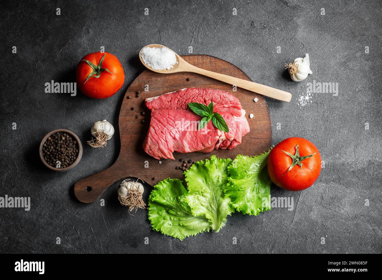 Bistecca di manzo cruda su tagliere su fondo rustico con foglie di menta, peperoni e lattuga, concetto di macelleria Foto Stock