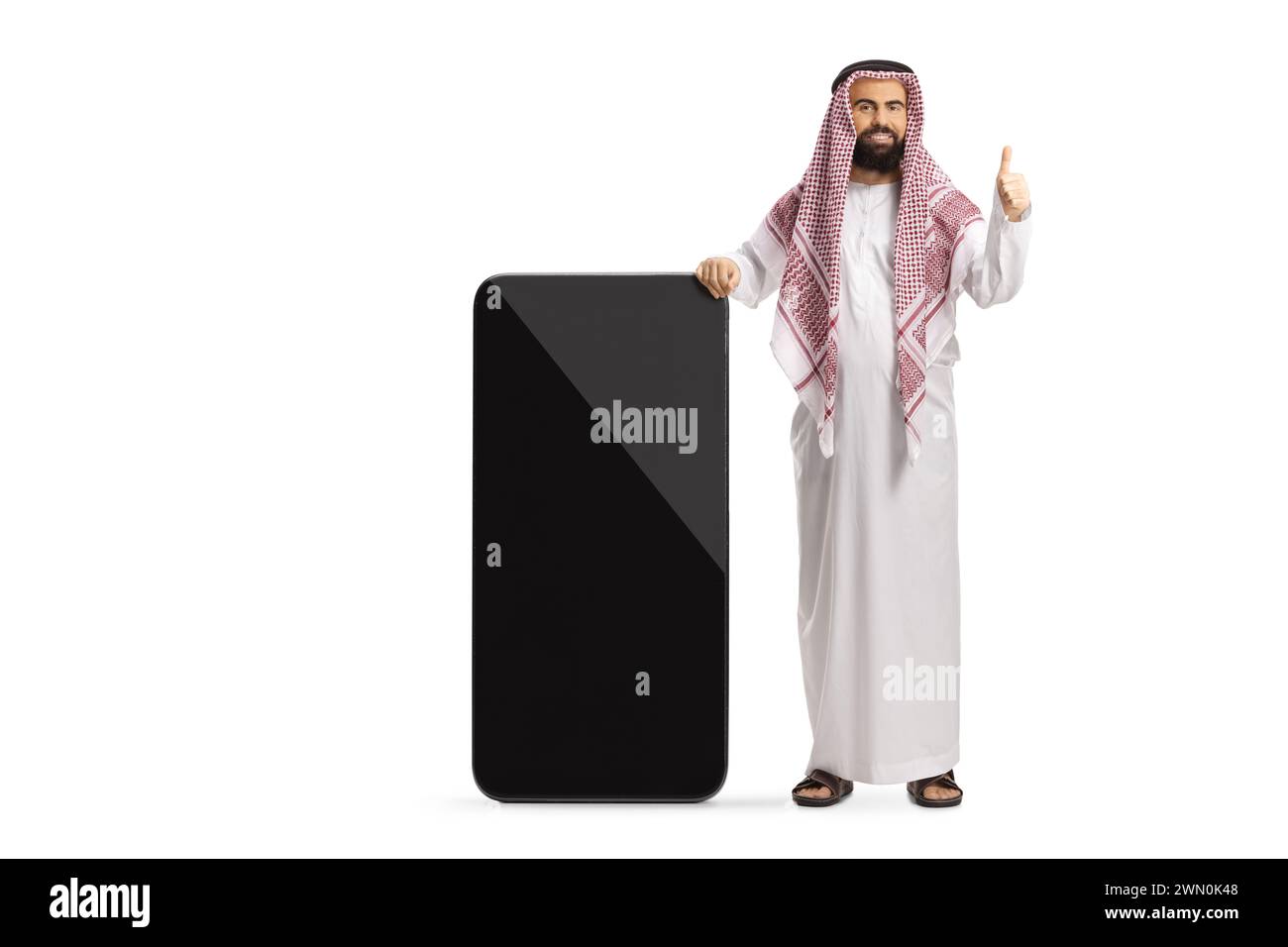 Uomo arabo saudita in abiti etnici appoggiato su un grande telefono cellulare e con gesti su un pollice isolato su sfondo bianco Foto Stock