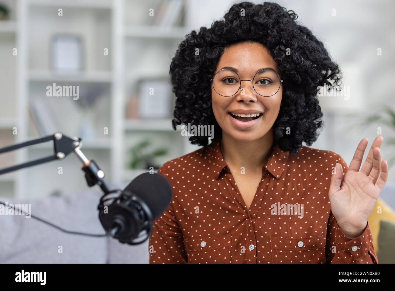 Una giovane donna afroamericana sta registrando una trasmissione online, insegnando da remoto, conversando in uno studio domestico e andando dal vivo. Si siede sul divano di fronte alla telecamera e saluta con un sorriso. Foto Stock