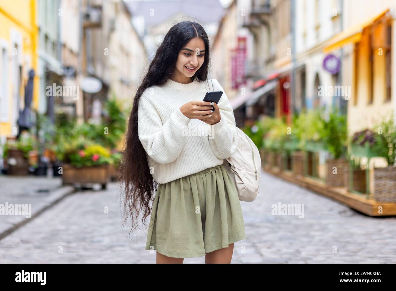 Una giovane ragazza indiana con un maglione bianco e uno zaino è in piedi per le strade della città e usa il telefono, digita un messaggio, effettua una chiamata. Foto Stock