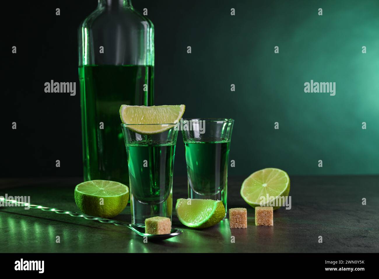 Absinthe in bicchieri da shot, cucchiaio, cubetti di zucchero di canna e lime su un tavolo testurizzato grigio su sfondo verde, spazio per il testo. Bevanda alcolica Foto Stock