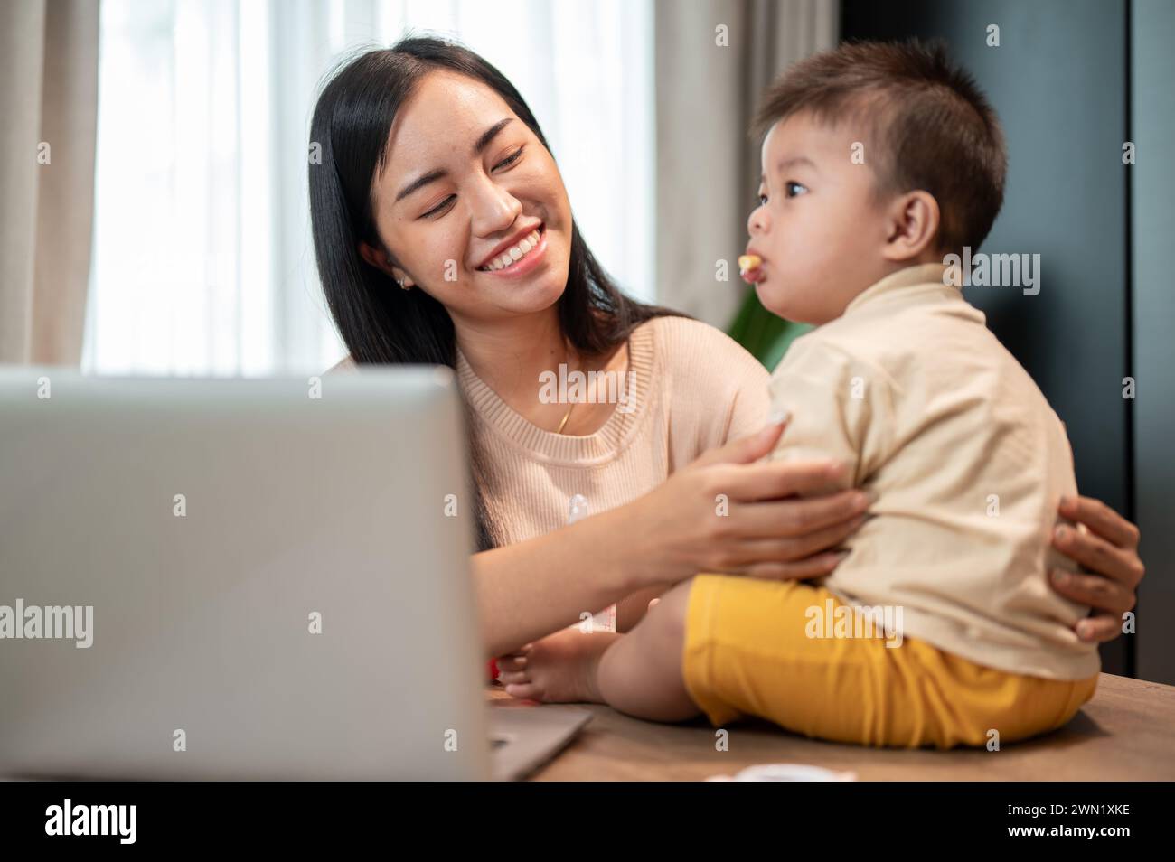 Una mamma asiatica felice sta giocando e nutrendo il suo bambino con spuntini mentre lavora nel suo ufficio domestico. lavora da casa e dai concetti di vita della mamma Foto Stock