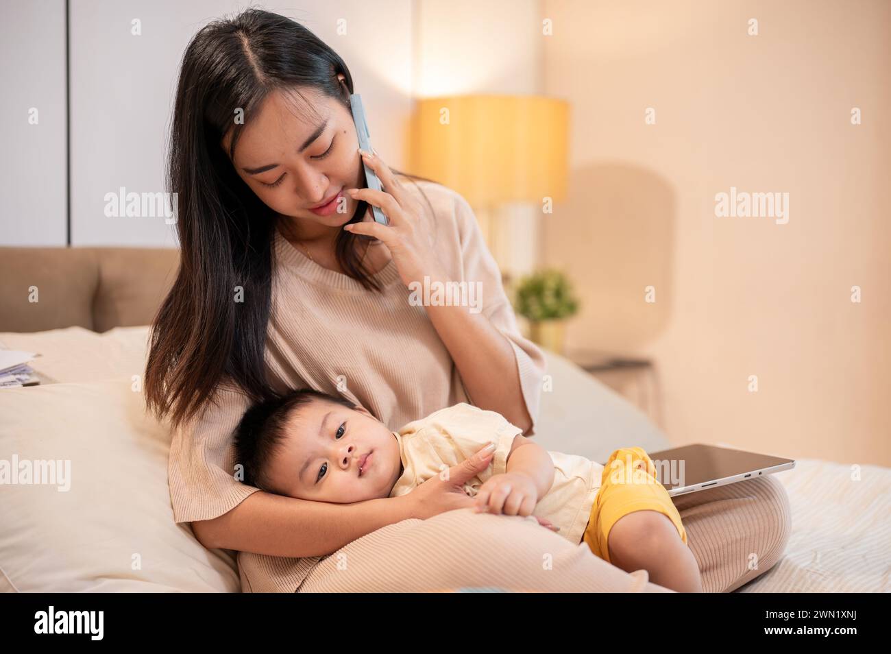 Una mamma asiatica premurosa sta mettendo il suo bambino a dormire tra le braccia mentre parla al telefono con qualcuno. pisolino e buonanotte, maternità e.. Foto Stock