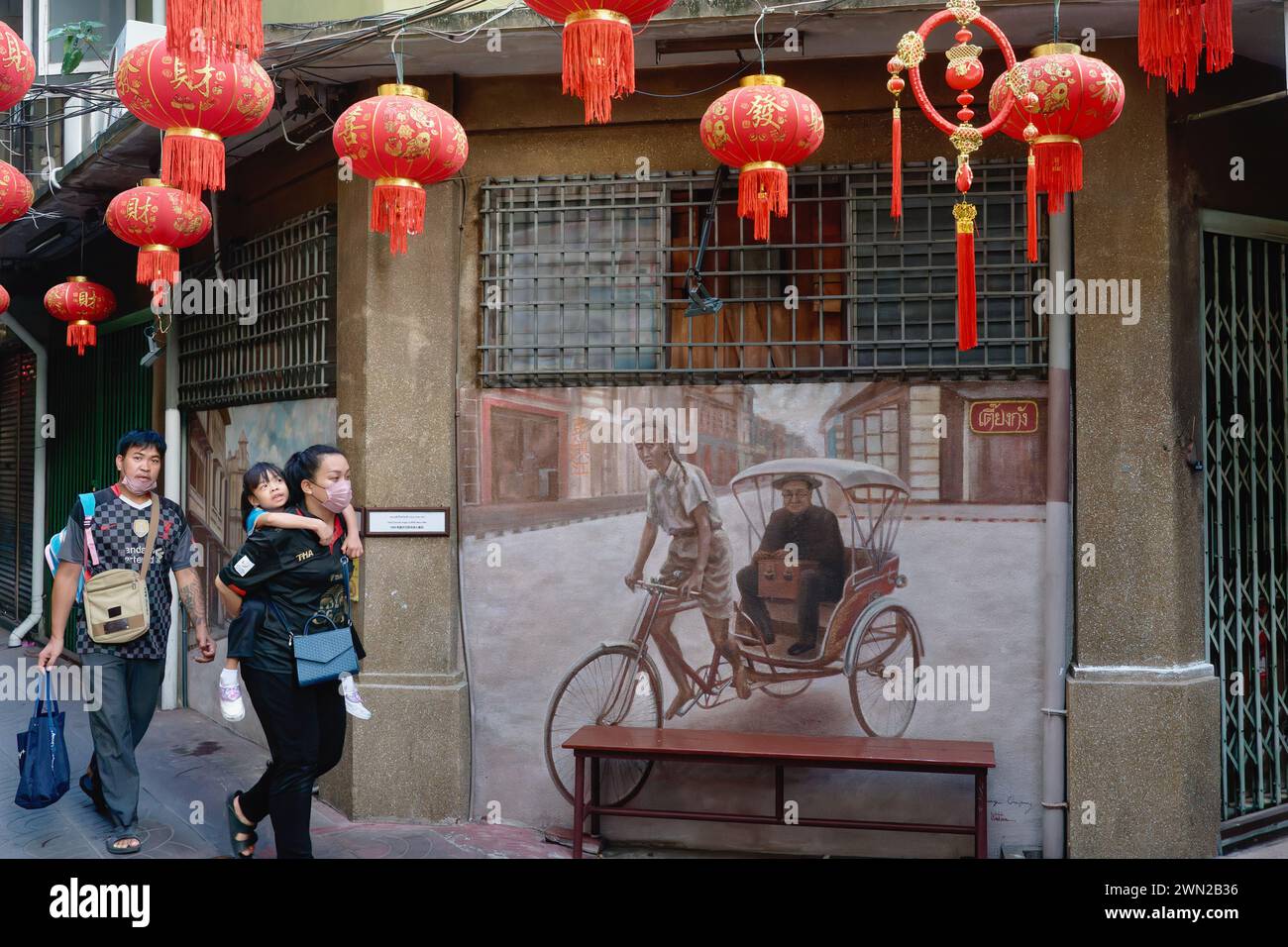 Una famiglia thailandese passa attraverso una stretta corsia a Chinatown, Bangkok, Thailandia, oltre un murale che raffigura un triciclo (trishaw) autista di un tempo passato Foto Stock