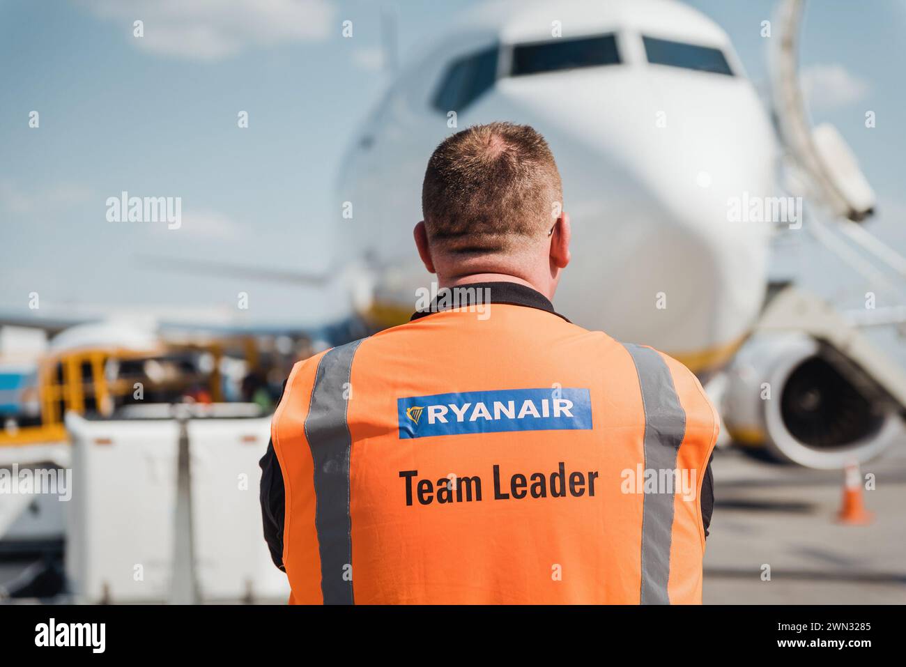 Ryanair team leader e Boeing 737 (EI-FZD) in una giornata di sole. Man in gilet arancione supervisiona l'assistenza a terra di un aeromobile all'arrivo Foto Stock