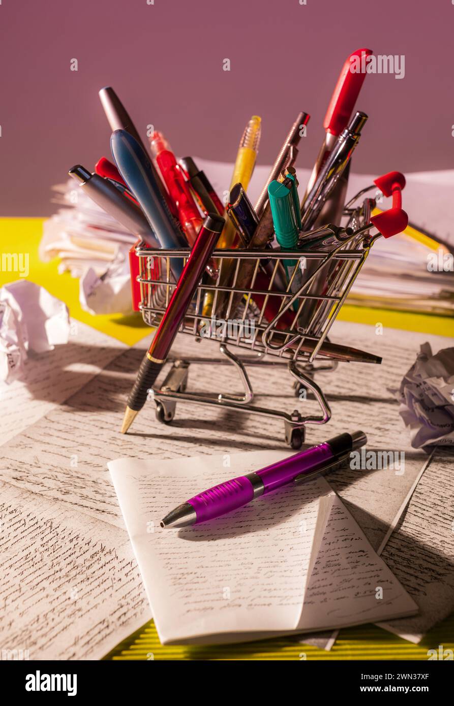 Un carrello della spesa in miniatura pieno di penne sulla scrivania di uno scrittore ingombrante, circondato da note scritte a mano e carte in una luce calda. Foto Stock