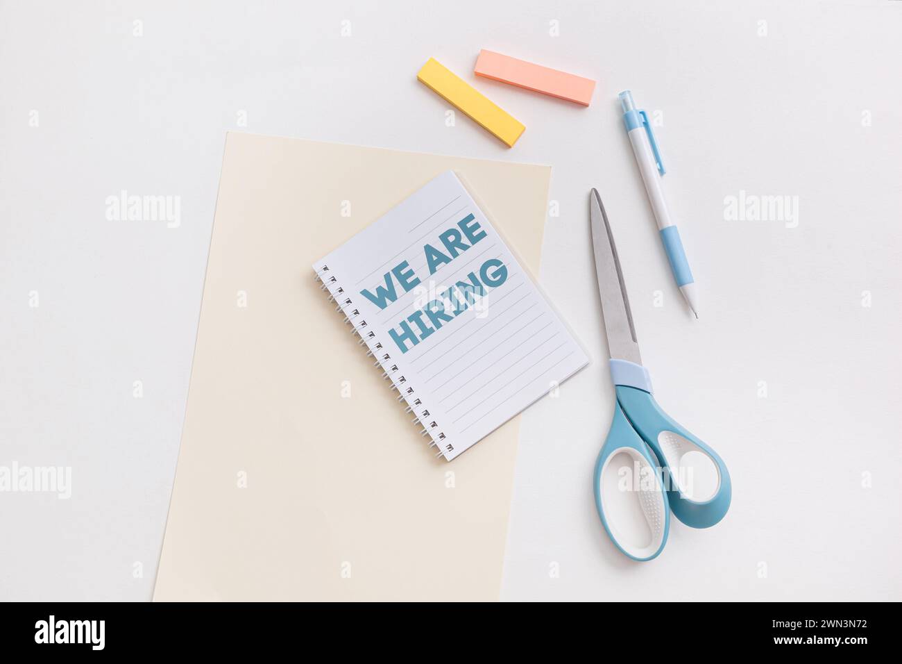 Una scrivania bianca con scritto We Are Hiring, con un quaderno blu di forbici, penna, evidenziatori, che simboleggia le opportunità di lavoro e. Foto Stock