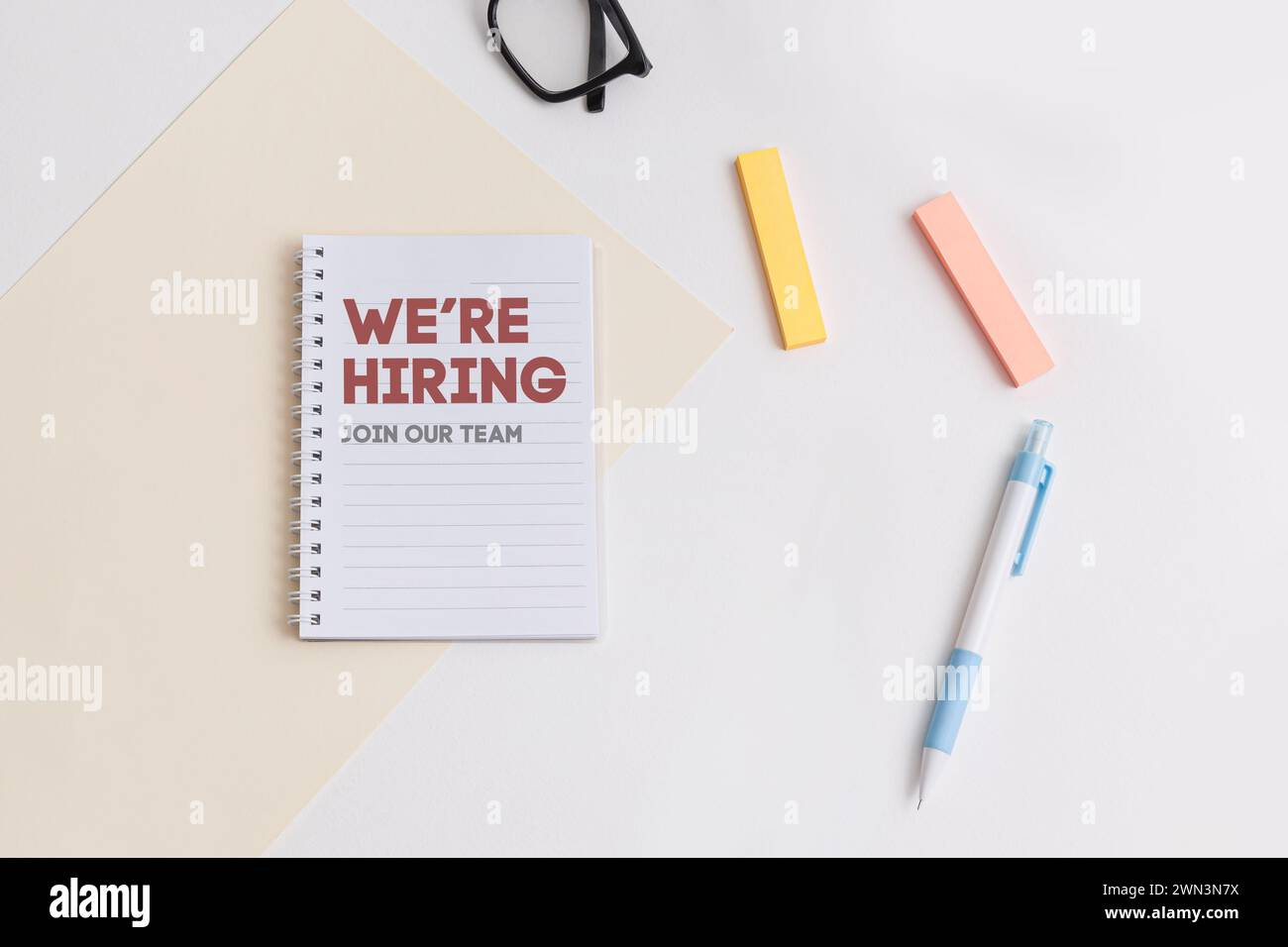 Una scrivania bianca che mostra un notebook con scritto We Are Hiring, penna blu, evidenziatori, simboleggia le opportunità di lavoro e l'ambiente di lavoro dell'ufficio Foto Stock