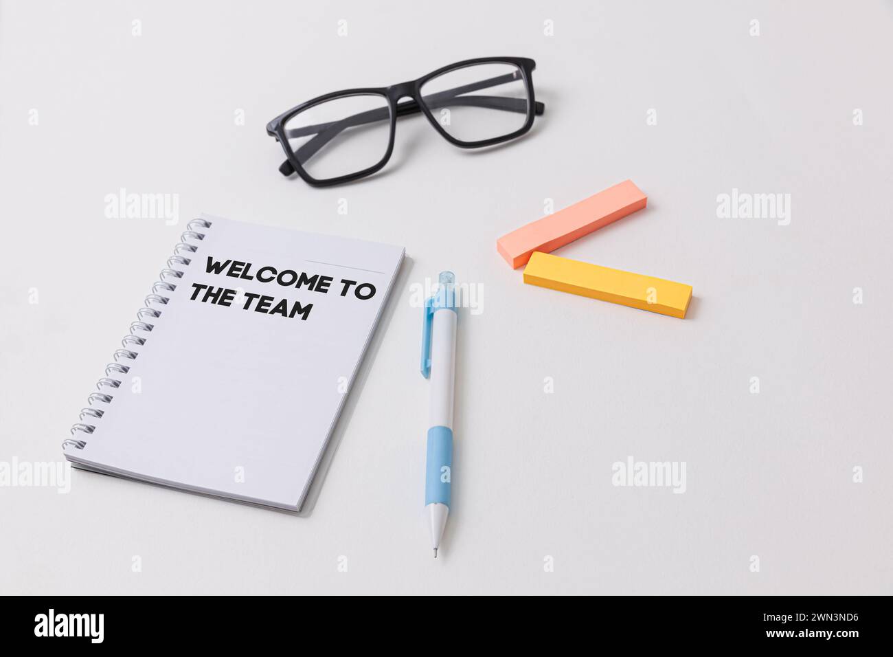 Una scrivania bianca che mostra un notebook con scritto benvenuto al team, penna blu, occhiali neri, evidenziatori, che simboleggia l'opportunità di lavoro e Foto Stock