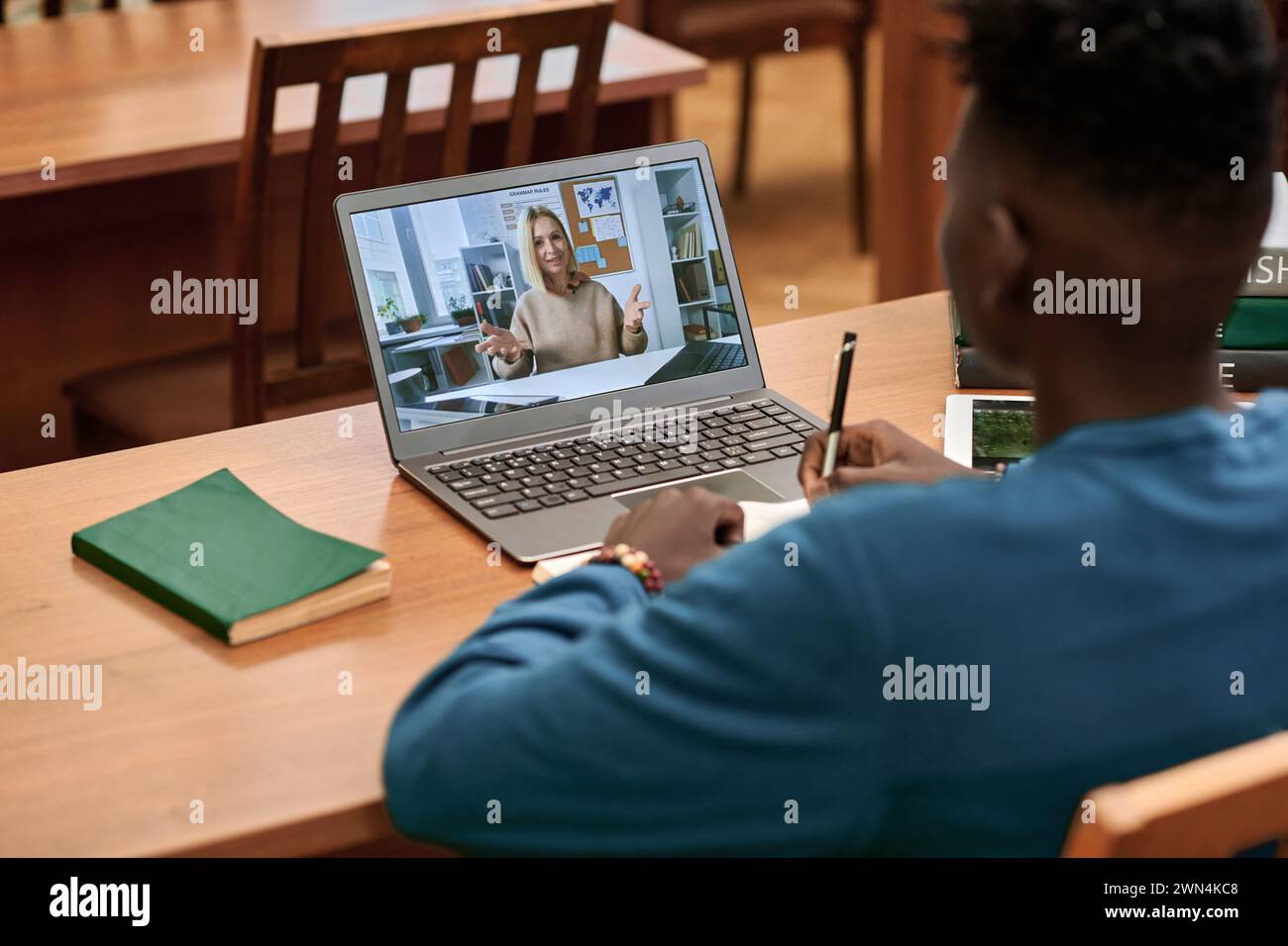 Vista posteriore del giovane uomo nero che guarda video lezioni nella biblioteca scolastica e utilizza un computer portatile con l'insegnante che parla sullo schermo Foto Stock