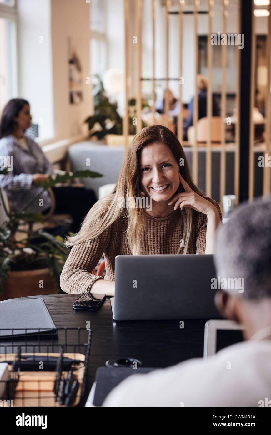 Ritratto di una donna d'affari sorridente seduta con un computer portatile alla scrivania in ufficio Foto Stock