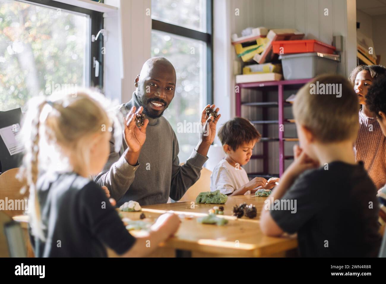 Insegnante sorridente che tiene in mano l'argilla mentre si siede con i bambini in classe in età prescolare Foto Stock