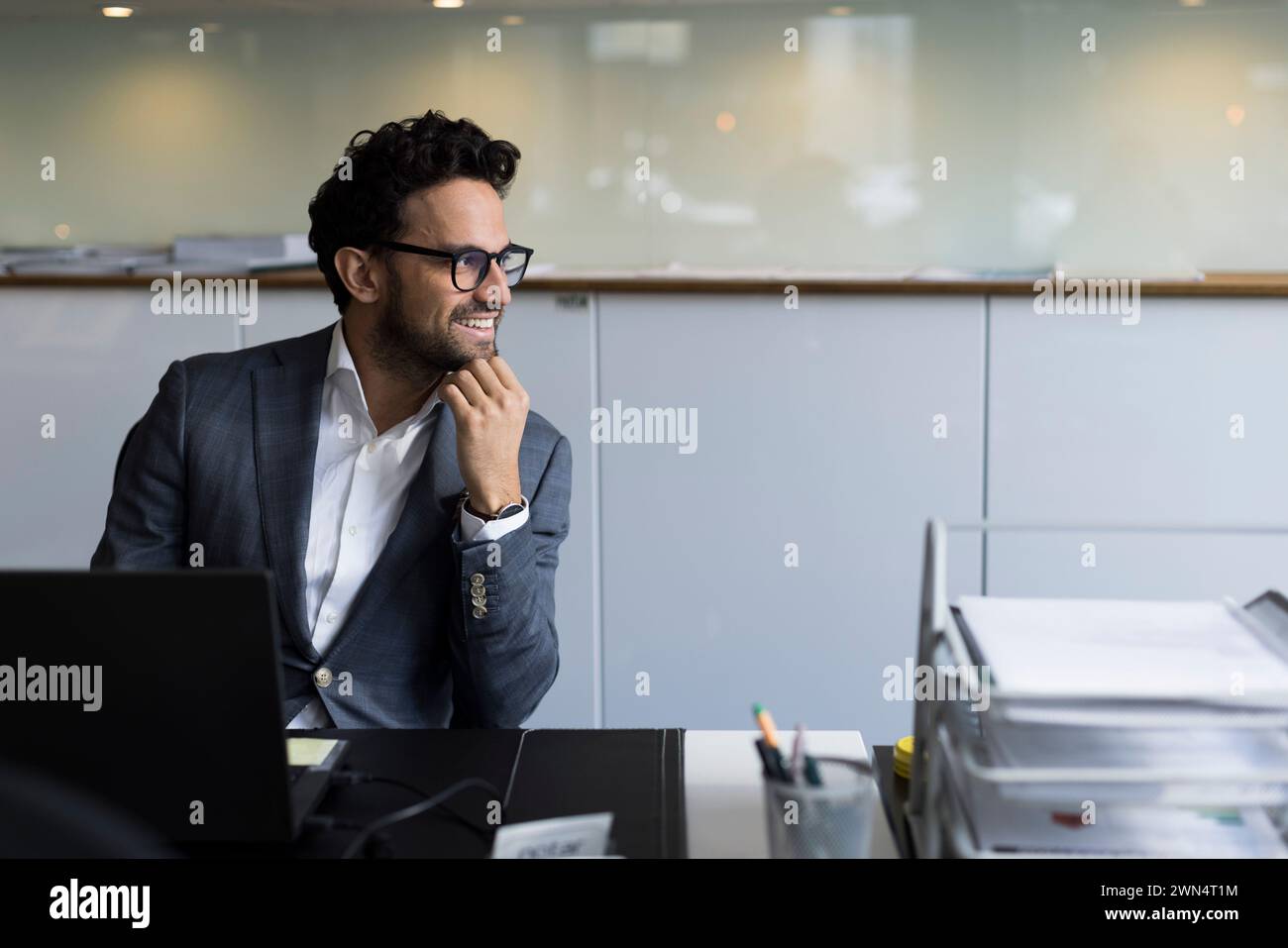 Un agente immobiliare sorridente che guarda lontano mentre si siede con la mano sul mento in ufficio Foto Stock