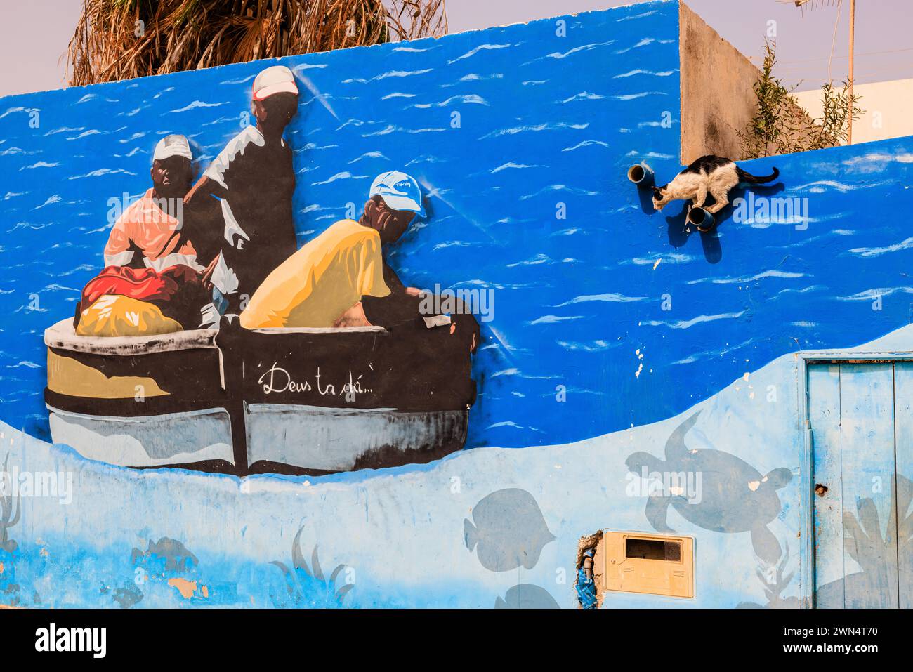 un gatto arroccato su un muro sembra prepararsi a saltare su una barca da pesca che è davvero un murale dipinto con colori vivaci su un muro a palmeira capo verde Foto Stock