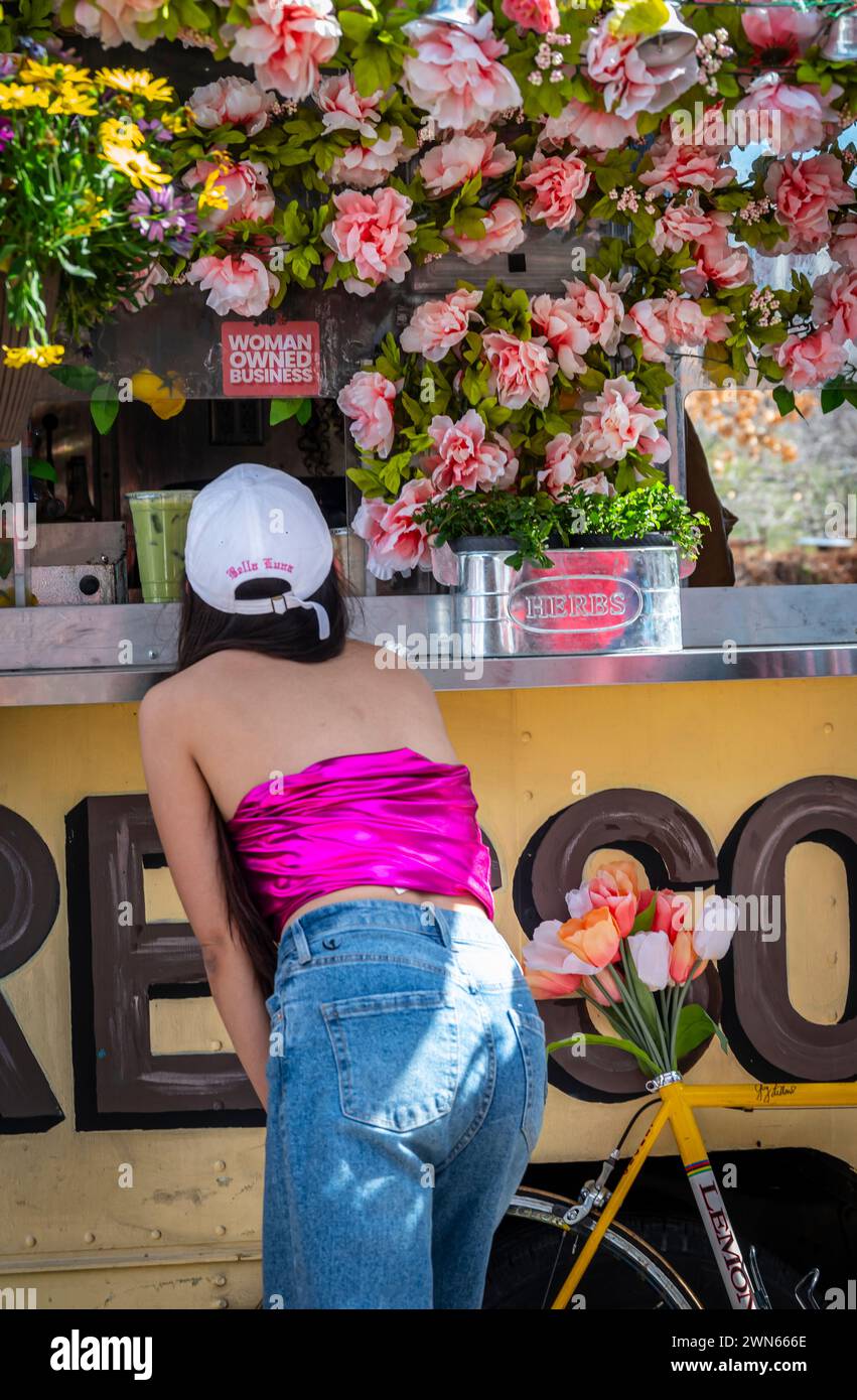 Vista posteriore di una donna in bandeau rosa caldo che ordina il succo dal food truck con un tema floreale e un cartello che recita UN'AZIENDA DI PROPRIETÀ DI UNA DONNA Foto Stock