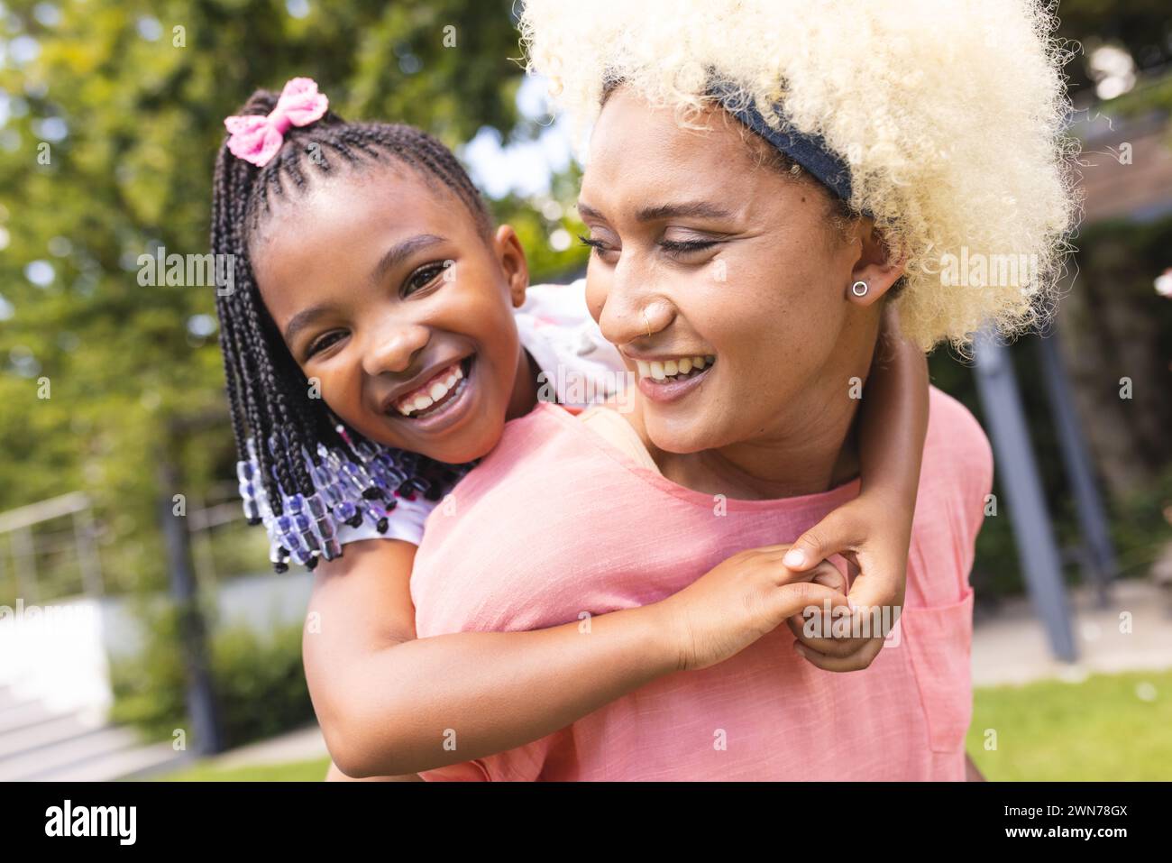 Figlia afro-americana con trecce e un fiocco sorride mentre fa il piggyback su una giovane madre birazziale Foto Stock