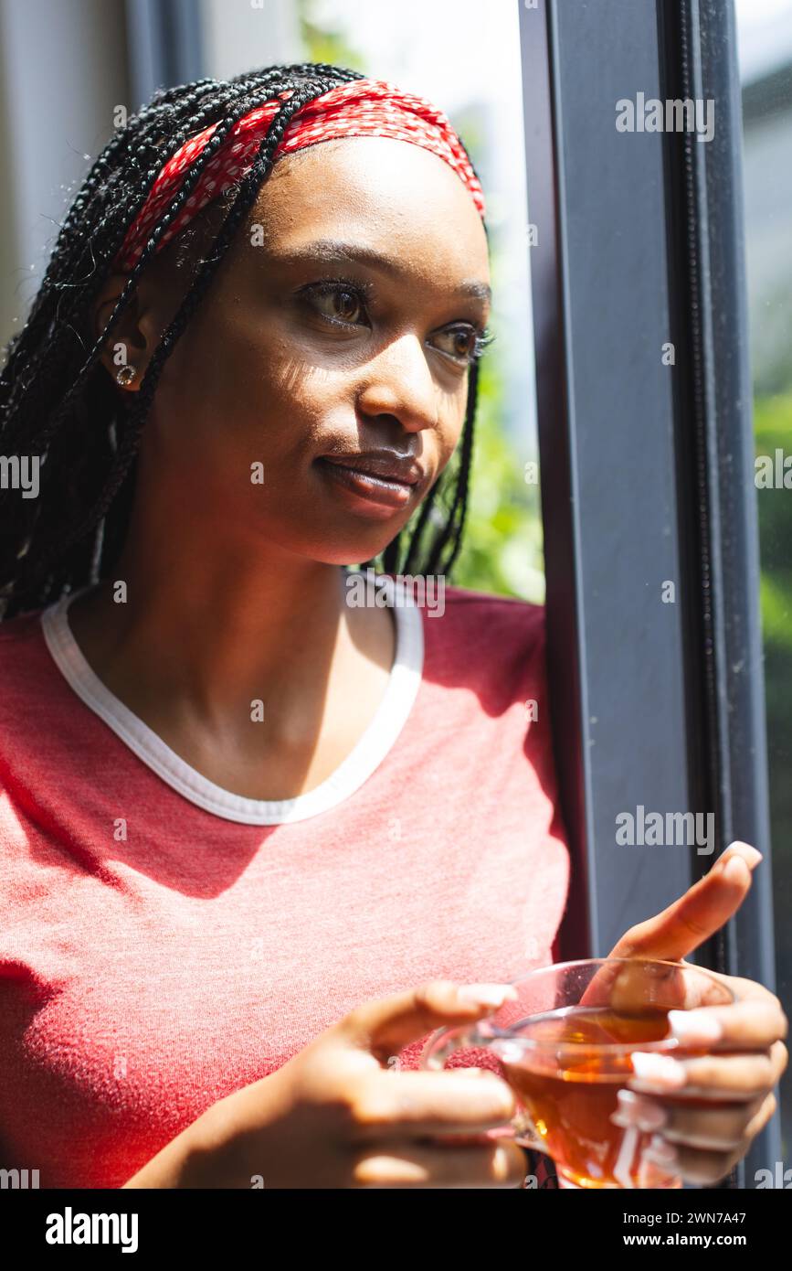 Una giovane donna afroamericana guarda fuori dalla finestra, tenendo una tazza di tè Foto Stock
