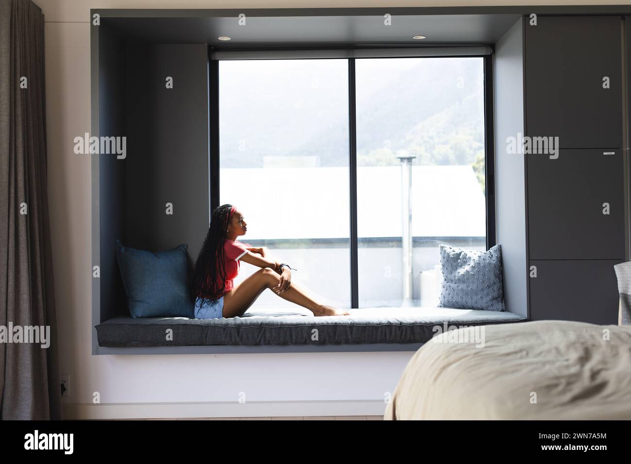Una giovane donna afro-americana siede accanto a una grande finestra, guardando fuori con spazio per copiare Foto Stock