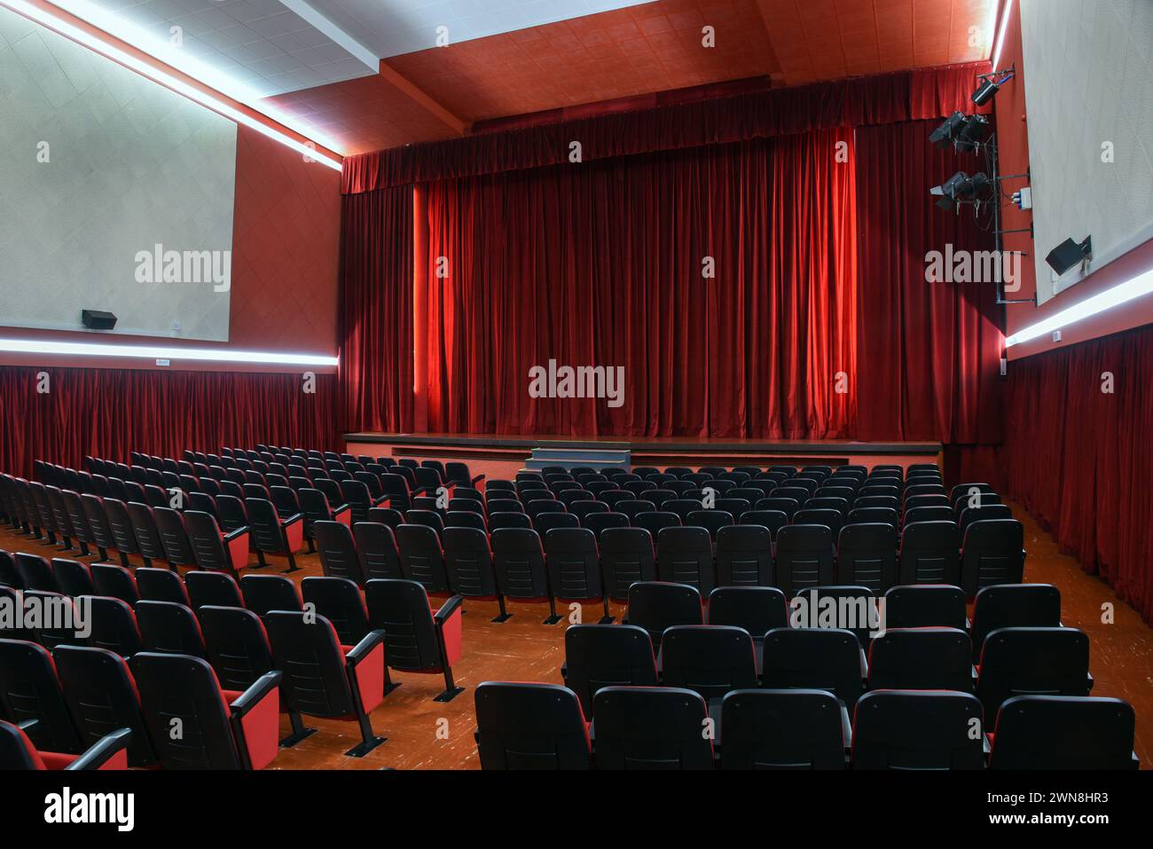 Interno di un teatro vuoto con file di comode poltrone sotto luci illuminanti, mentre tende rosse luminose appese lungo le pareti e dal palco ce Foto Stock