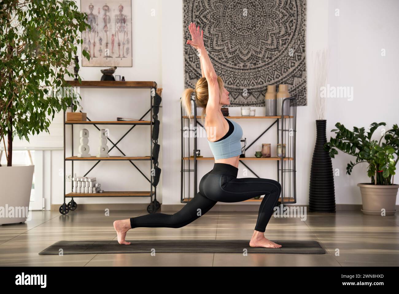 Corpo completo di donna flessibile in abbigliamento sportivo che pratica Anjaneyasana posa sul tappeto durante l'allenamento di yoga in un moderno studio decorato con piante in vaso e ta Foto Stock