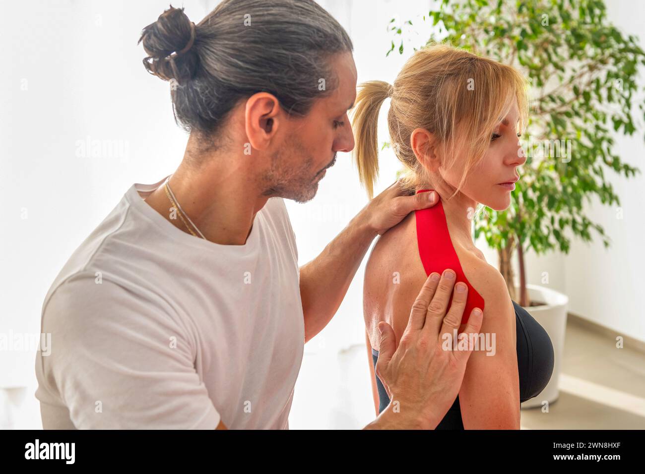 Massaggiatore esperto che tratta la spalla della signora ferita con kinesiotape rossa durante l'allenamento di yoga in sala fitness contro una pianta in vaso vicino alla parete bianca Foto Stock