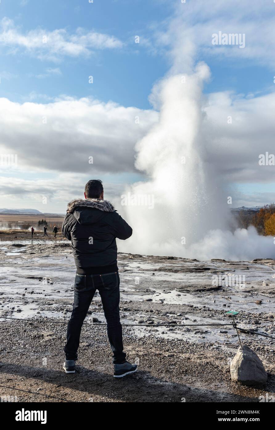 Vista posteriore di un anonimo viaggiatore maschile che guarda lontano mentre si trova su un terreno vulcanico roccioso con acqua alla luce del giorno e ammira il vapore bianco Foto Stock