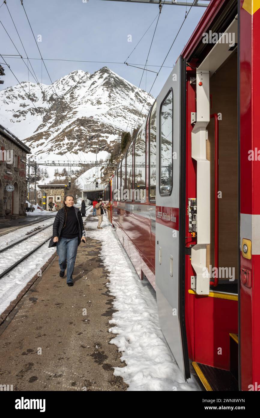 Una camminata femminile lungo il treno Bernina Express nelle Alpi svizzere, con montagne innevate sullo sfondo, terreno parzialmente coperto di neve Foto Stock