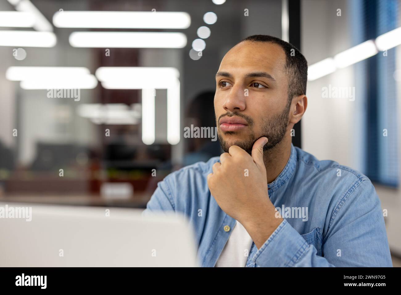 Un giovane professionista contemplativo in un ambiente d'ufficio ben illuminato, colto in un momento di attenzione mentre si guarda lontano dallo schermo del suo notebook. La sua espressione trasmette concentrazione e pensiero profondo. Foto Stock