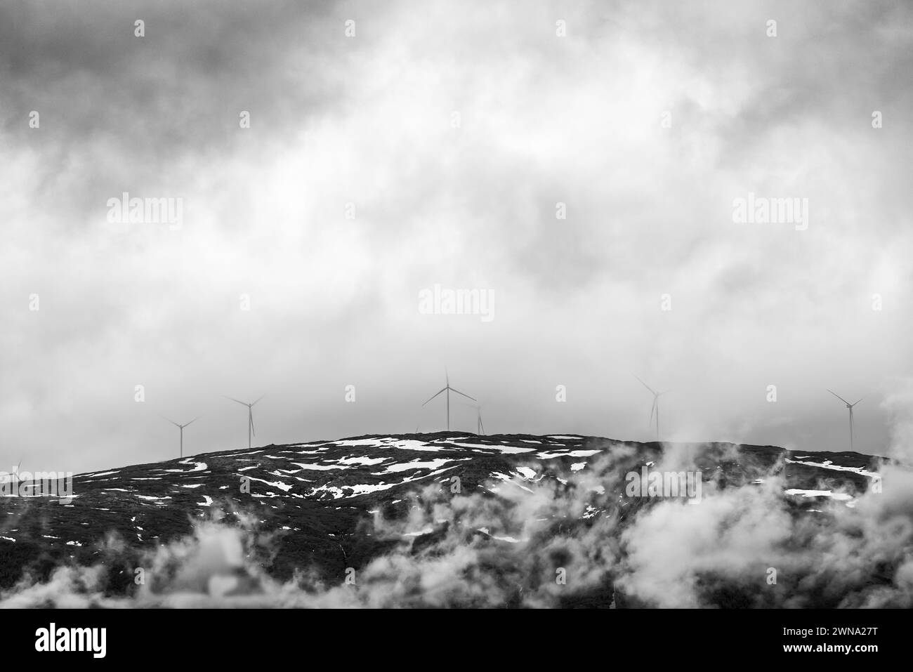 Le turbine eoliche si allineano lungo una cresta montuosa ricoperta di neve in Norvegia, parzialmente velata da nuvole basse in un paesaggio drammatico in bianco e nero. Foto Stock
