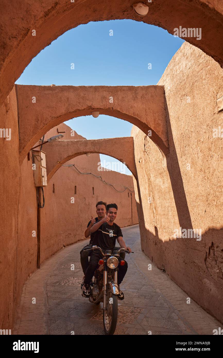 Due giovani uomini iraniani sorridono e mostrano il pollice in alto mentre guidano in moto in una strada stretta nello storico quartiere Fahadan di Yazd, Iran. Foto Stock