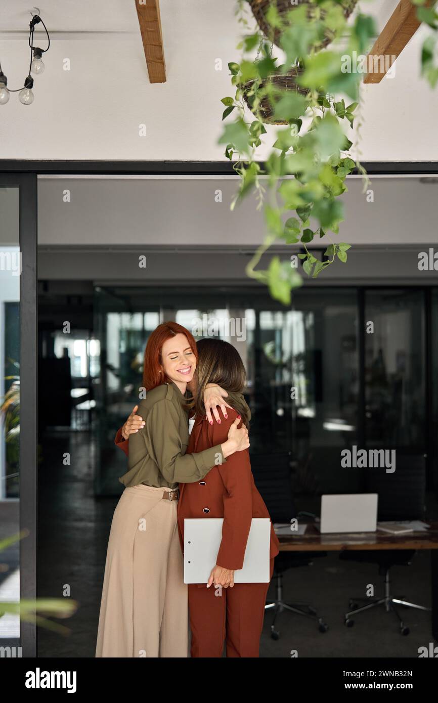 Due donne d'affari felici in piedi al lavoro in ufficio abbracciato, verticale. Foto Stock
