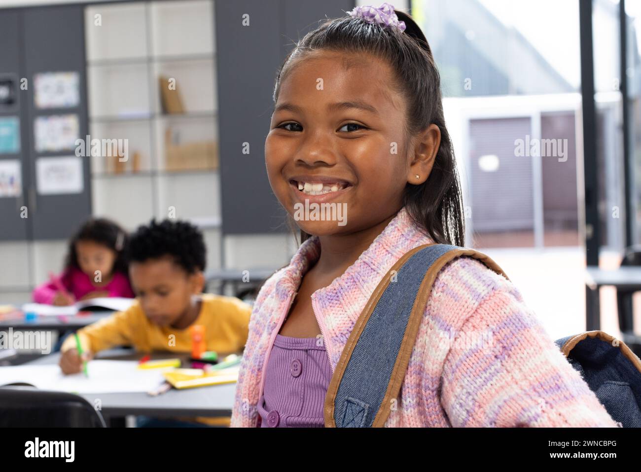 Una ragazza birazziale con un accessorio per capelli viola sorride, indossando uno zaino in una classe scolastica Foto Stock