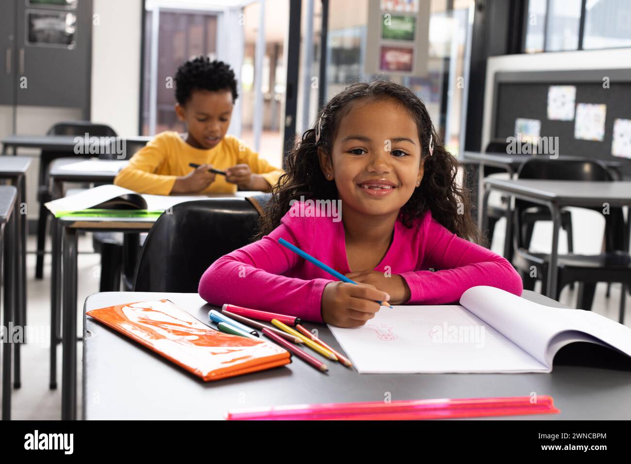 Una ragazza birazziale con i capelli ricci sorride mentre disegna in una classe scolastica, indossando un top rosa Foto Stock