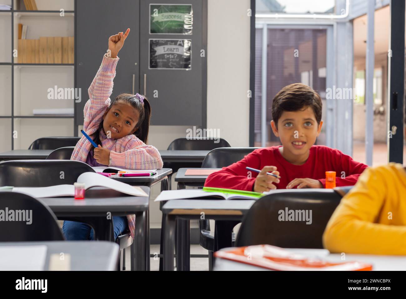 Una ragazza birazziale con una camicia rosa a quadri alza la mano con impazienza in una classe scolastica Foto Stock