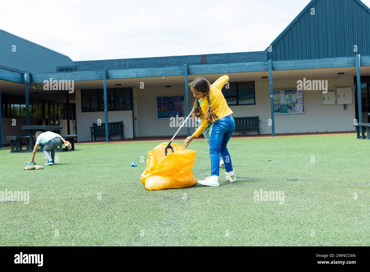Una ragazza birazziale in cima gialla partecipa a una pulizia, raccogliendo spazzatura in un sacchetto giallo Foto Stock
