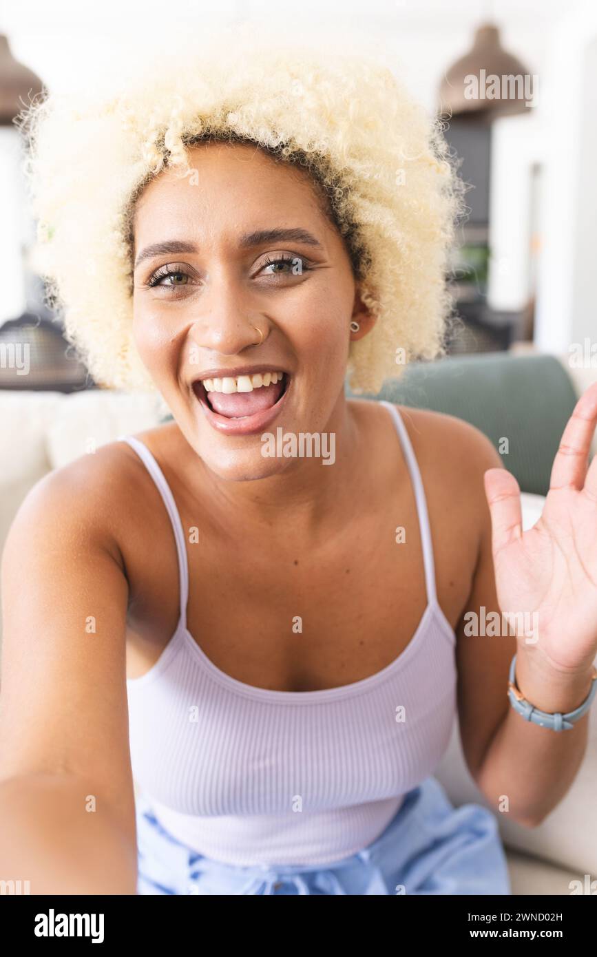 Giovane donna birazziale con capelli biondi ricci sorride gioiosamente, salutando la telecamera durante una videochiamata Foto Stock